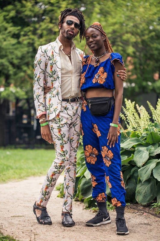 A man and woman posing, he is wearing a white suit with parrots on it while she is wearing a dark bl...