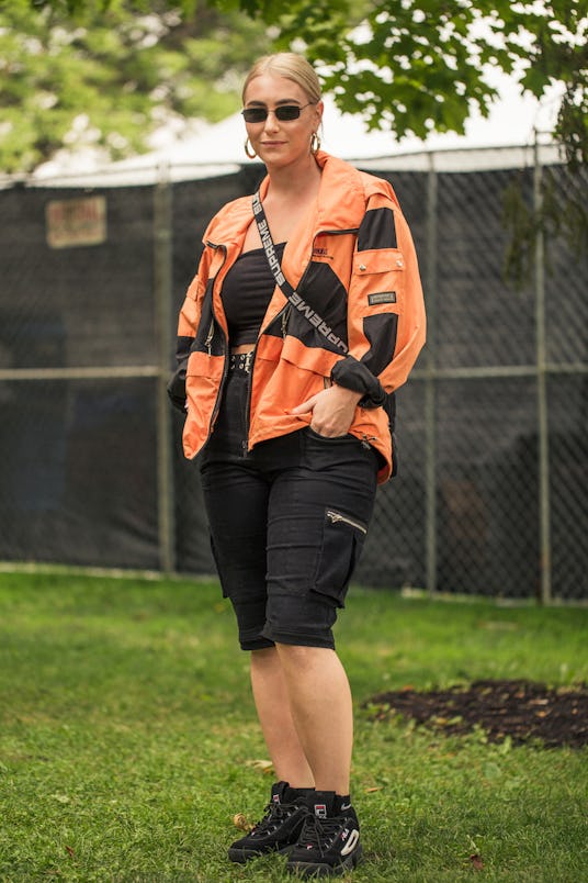 A girl in an athleisure outfit: an orange wind coat, black top, black cargo knee-length shorts, blac...