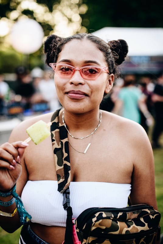 A woman with space buns, wearing a white bandeau top, pink geometric sunglasses and a camo bag at Go...