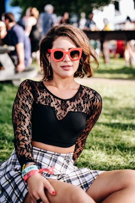 Maya Klausner in a black lace top, a plaid skirt and large orange sunglasses, sitting on the grass a...