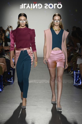 Two models walking in a burgundy top and green pants, and pink shorts with a green top and pink vest...
