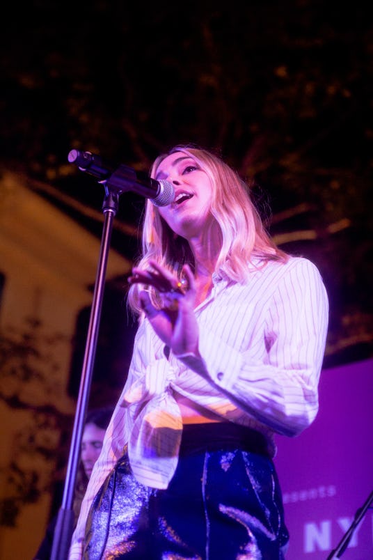 Katelyn Tarver wearing a white tied-up top singing into the microphone whilst playing a brown guitar
