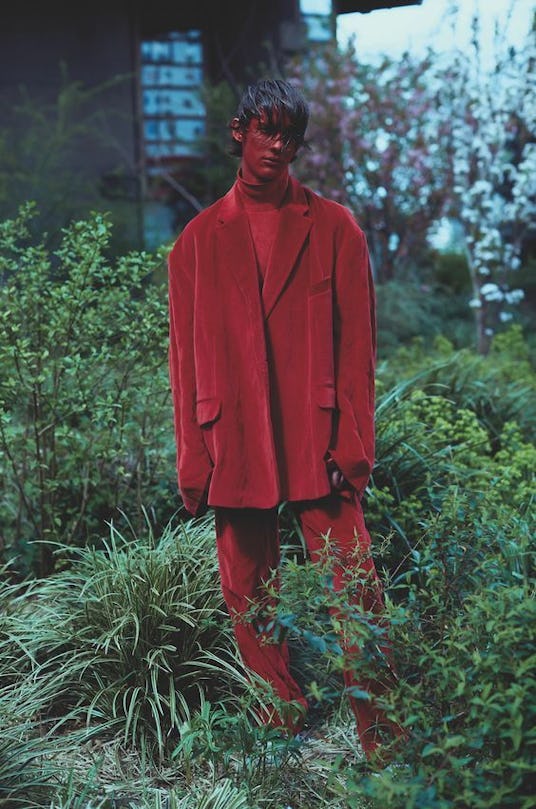 A man with red paint over his face posing while wearing a red blazer, red sweater, and red pants