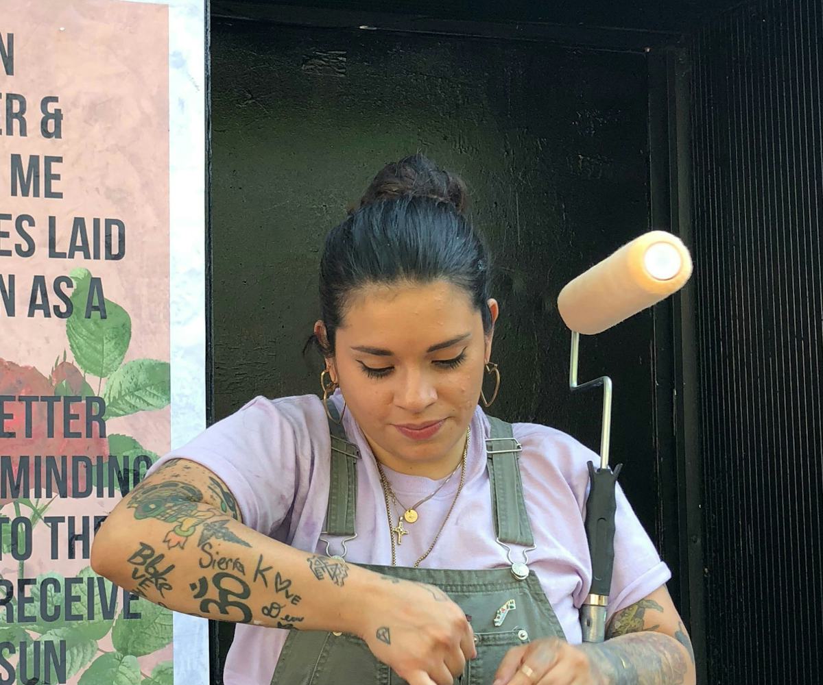 Johanna Toruño, in grey overalls with a paint roller, standing in front of a wall poster with her me...