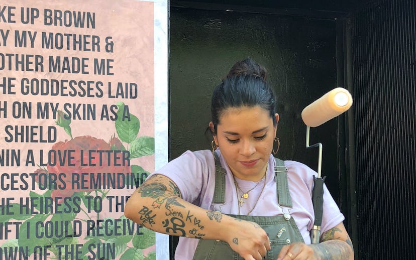 Johanna Toruño, in grey overalls with a paint roller, standing in front of a wall poster with her me...