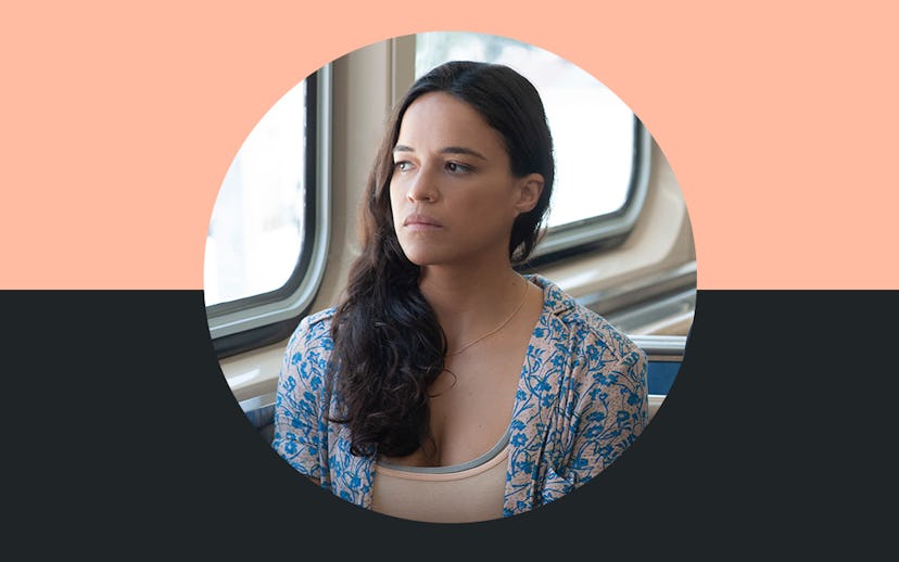 Michelle Rodriguez sitting in a train next to a window in a blue-beige floral jacket and beige top