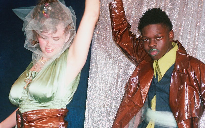 Couple looking like they are at an '80s prom, girl in a green dress and a brown leather belt, guy in...