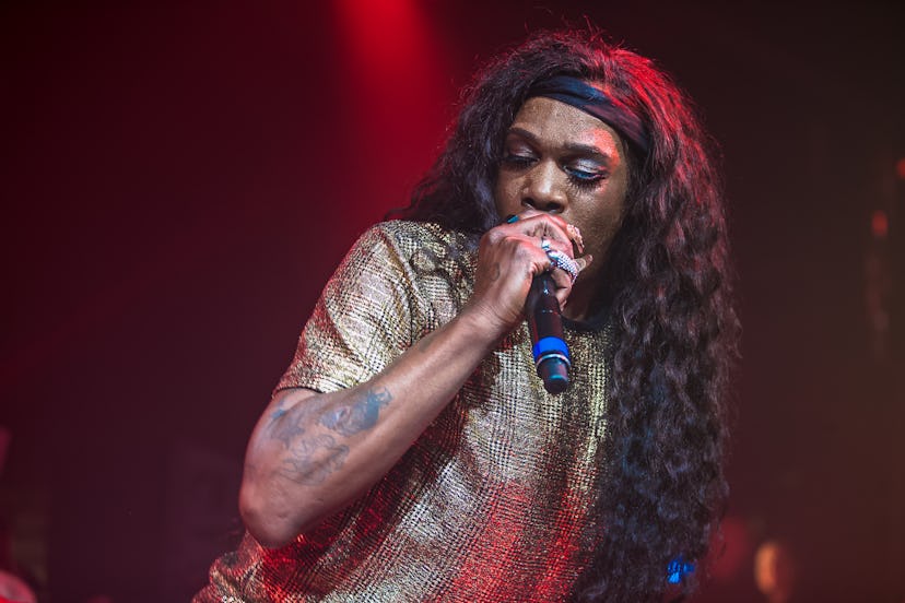 Big Freedia singing with a microphone in a golden printed short-sleeved shirt and black hairband