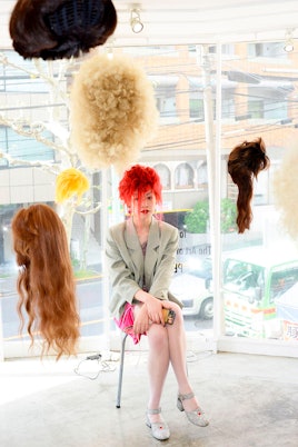 A woman sitting and wearing a red wig, surrounded by hanging wigs, at the Tomi Kono "Personas" Exhib...