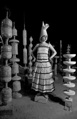 Flavia Da Rin standing on a brick while wearing a dress and a big headpiece surrounded by geometric ...