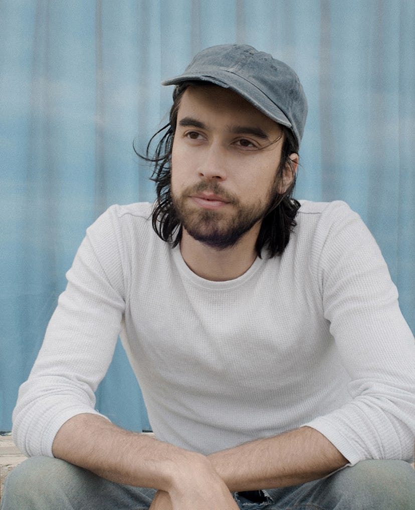 Alex G in a white long-sleeved shirt with light grey jeans and a grey denim cap