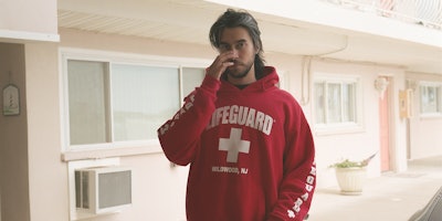Long-haired Alex G wearing a red hoodie with a text on it that says lifeguard.