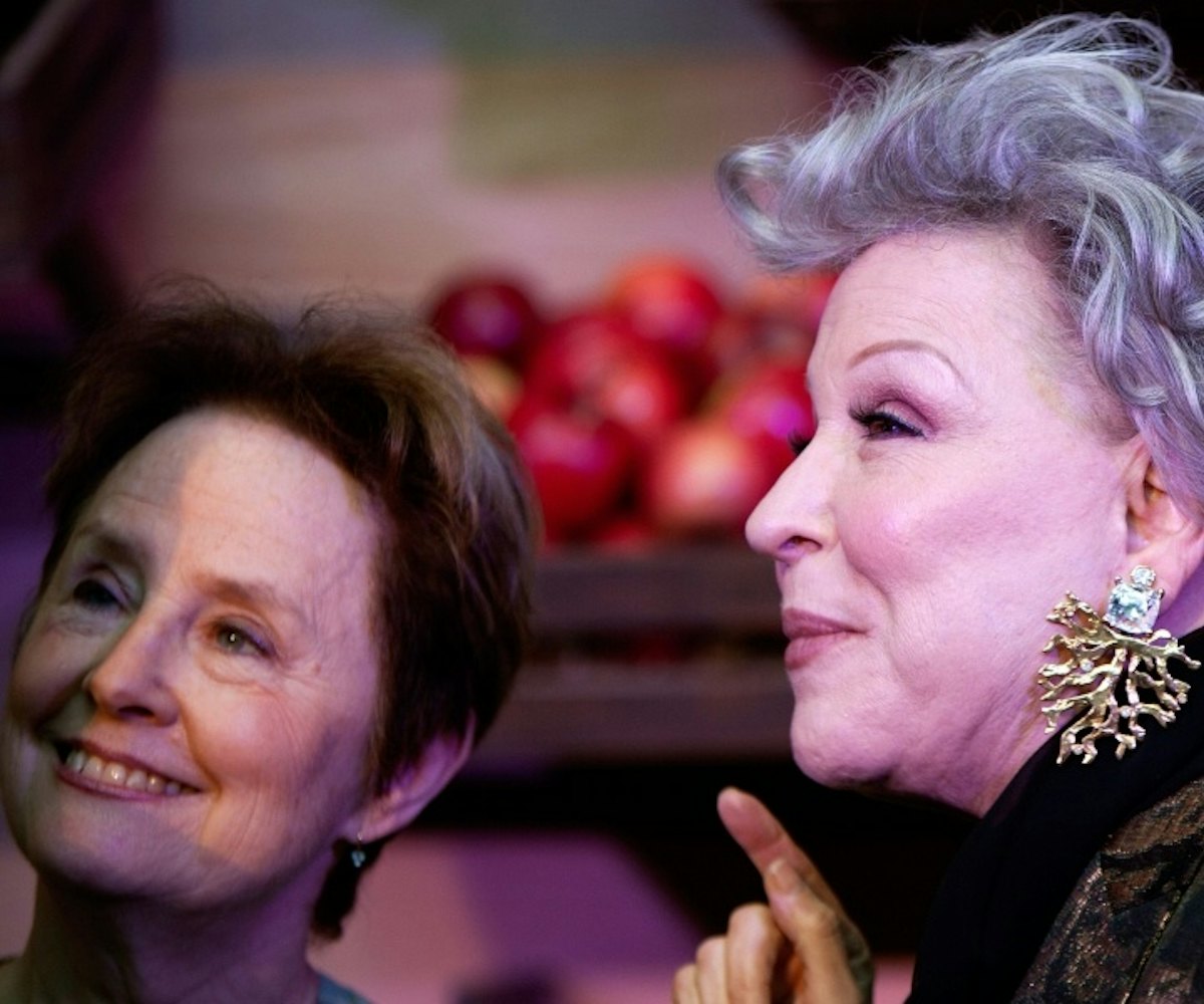 Bette Midler with diamond tree root earrings standing in front of a crate of apples