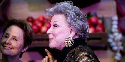 Bette Midler with diamond tree root earrings standing in front of a crate of apples
