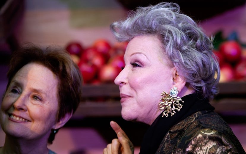 Bette Midler with diamond tree root earrings standing in front of a crate of apples