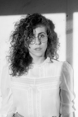 Ariana Reines in black and white wearing a white top and glasses looking at the camera
