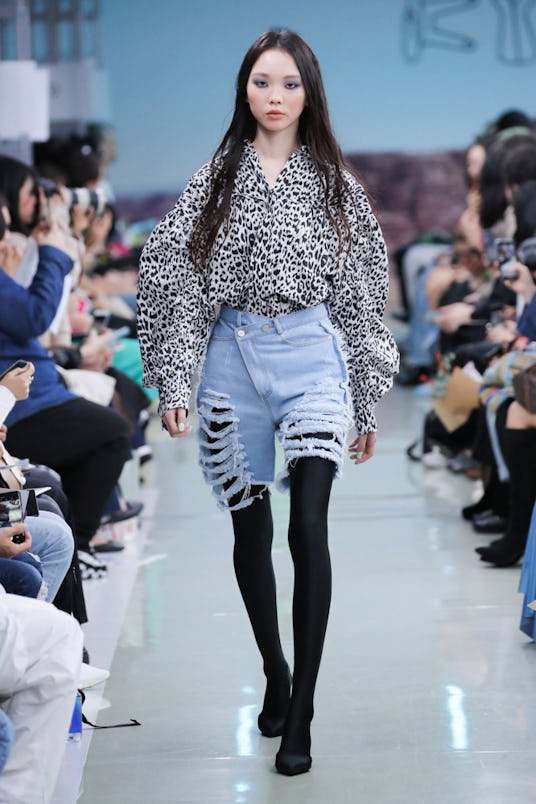 A female model walking on a runway while wearing denim shorts, black leggings, and a black and white...