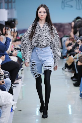 A female model walking on a runway while wearing denim shorts, black leggings, and a black and white...