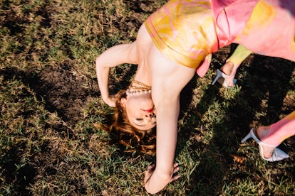 Jessica Barden performing a gymnastic bridge on the grass in a peach-yellow silk wrap-tie top