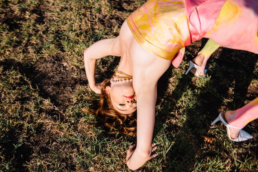 Jessica Barden performing a gymnastic bridge on the grass in a peach-yellow silk wrap-tie top