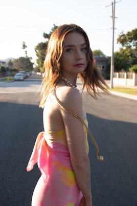 Jessica Barden on the street looking over her shoulder in a peach-yellow silk wrap-tie top and peach...