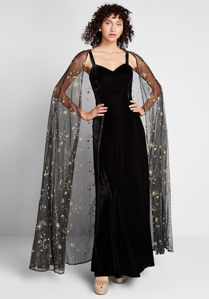 A model in Modcloth, Celestial Occasion Velvet Maxi Dress in black showing off a sheer cape with sta...