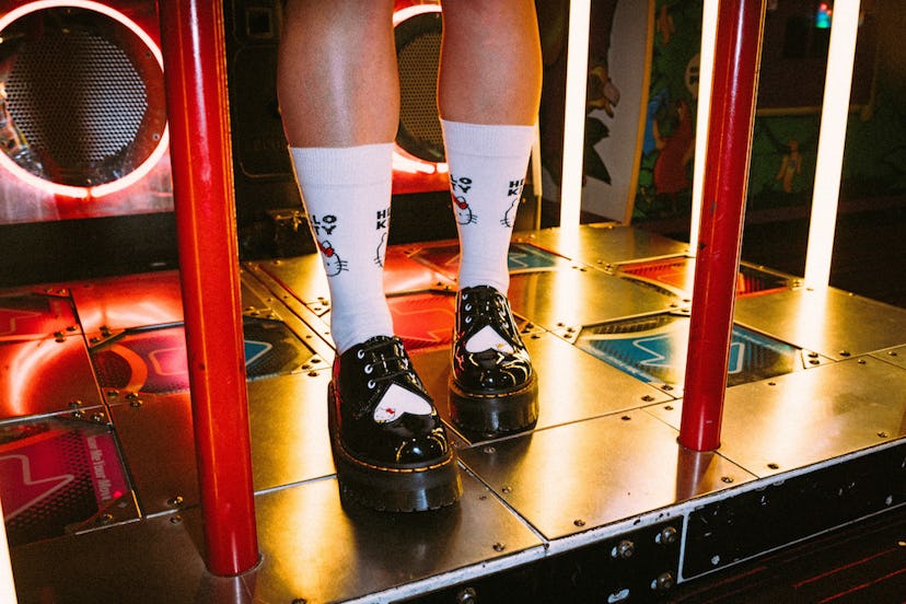 A person wearing black Dr. Martens 1461 Hello Kitty Loafers and white socks standing between two red...