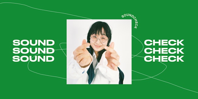Artist Yaeji in a white shirt doing heart signs gestures with her fingers with a green background wi...