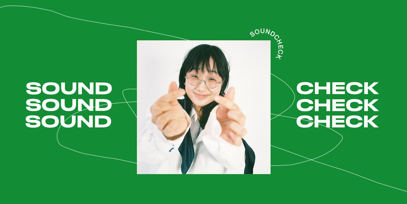 Artist Yaeji in a white shirt doing heart signs gestures with her fingers with a green background wi...