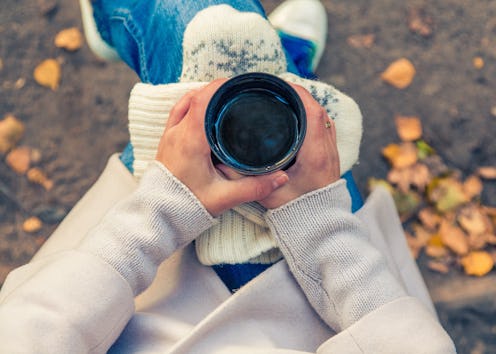 A woman holding a cup of coffee over her gloves on her knees while sitting in a park with autumn lea...