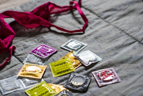 Many different condom packages on a bed next to a burgundy bra