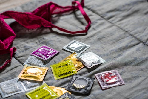 Many different condom packages on a bed next to a burgundy bra