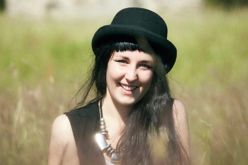 A woman sitting in tall grass with a black bowler hat smiling after running