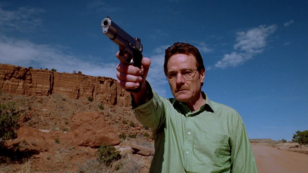 Walter White, holding a gun in front of his face, pointed towards something off-screen. Played by Bryan Cranston, the character is wearing a green shirt and glasses and standing in the New Mexico desert.