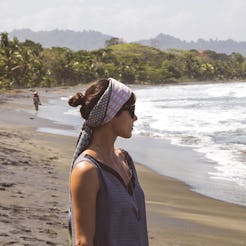 A woman wearing a headscarf and a grey shirt with a glowing, healthy skin standing on a beach next t...