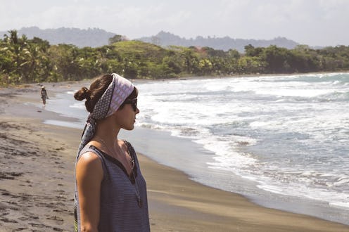A woman wearing a headscarf and a grey shirt with a glowing, healthy skin standing on a beach next t...