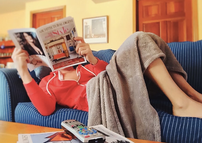 A woman lying on a couch covered with a blanket reading a newspaper