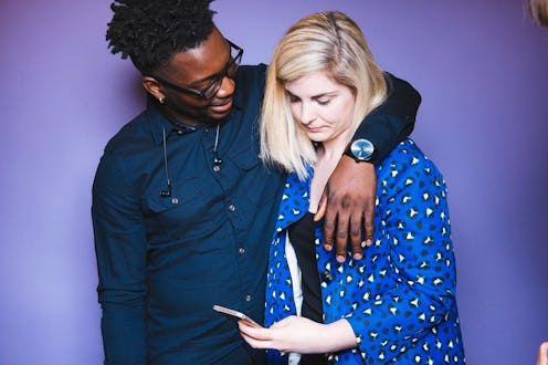 A man wearing glasses and a denim shirt hugging his partner with ADHD while she is looking at her ph...