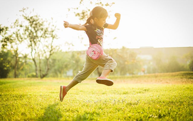 A girl with pigtails in a black T-shirt and gray pants, and a pink purse skipping through a sunny fi...