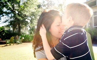 A boy in a dark blue T-shirt with thin white stripes kissing his brunette mother's forehead in the g...