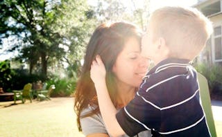 A boy in a dark blue T-shirt with thin white stripes kissing his brunette mother's forehead in the g...