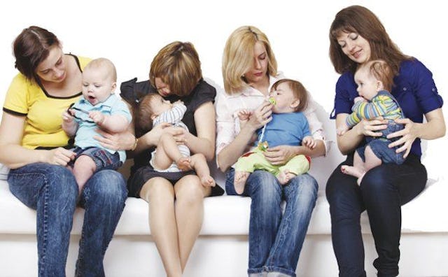 Four mom friends on a white couch holding their toddler and interacting with them.