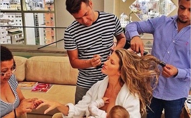 Gisele Bundchen in a white robe breastfeeding her baby while two men are doing her hair and a woman ...