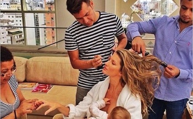 Gisele Bundchen in a white robe breastfeeding her baby while two men are doing her hair and a woman ...