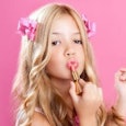 A blonde girl with pink accessories in her hair putting pink lipstick on her lips