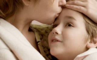 A close-up of a mother in a beige shirt with her child in her lap whose forehead she's kissing while...