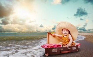 A baby in an orange dress and an oversized straw hat is sitting in a brown suitcase which is located...