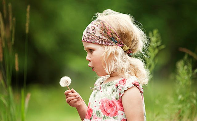 A young blonde girl blowing toward a dandelion while standing in nature.