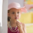 A small girl putting her mother's lipstick on while wearing a pink lady hat