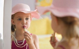 A girl putting her mother's lipstick on while wearing a pink hat, pink dress and a long pearl neckla...